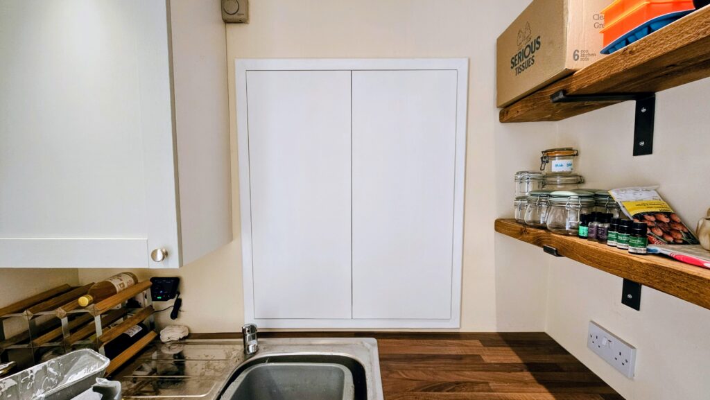 Wide-angle view of custom boiler cupboard doors installed in a kitchen in Glossop, matching the surrounding cabinetry and shelving.